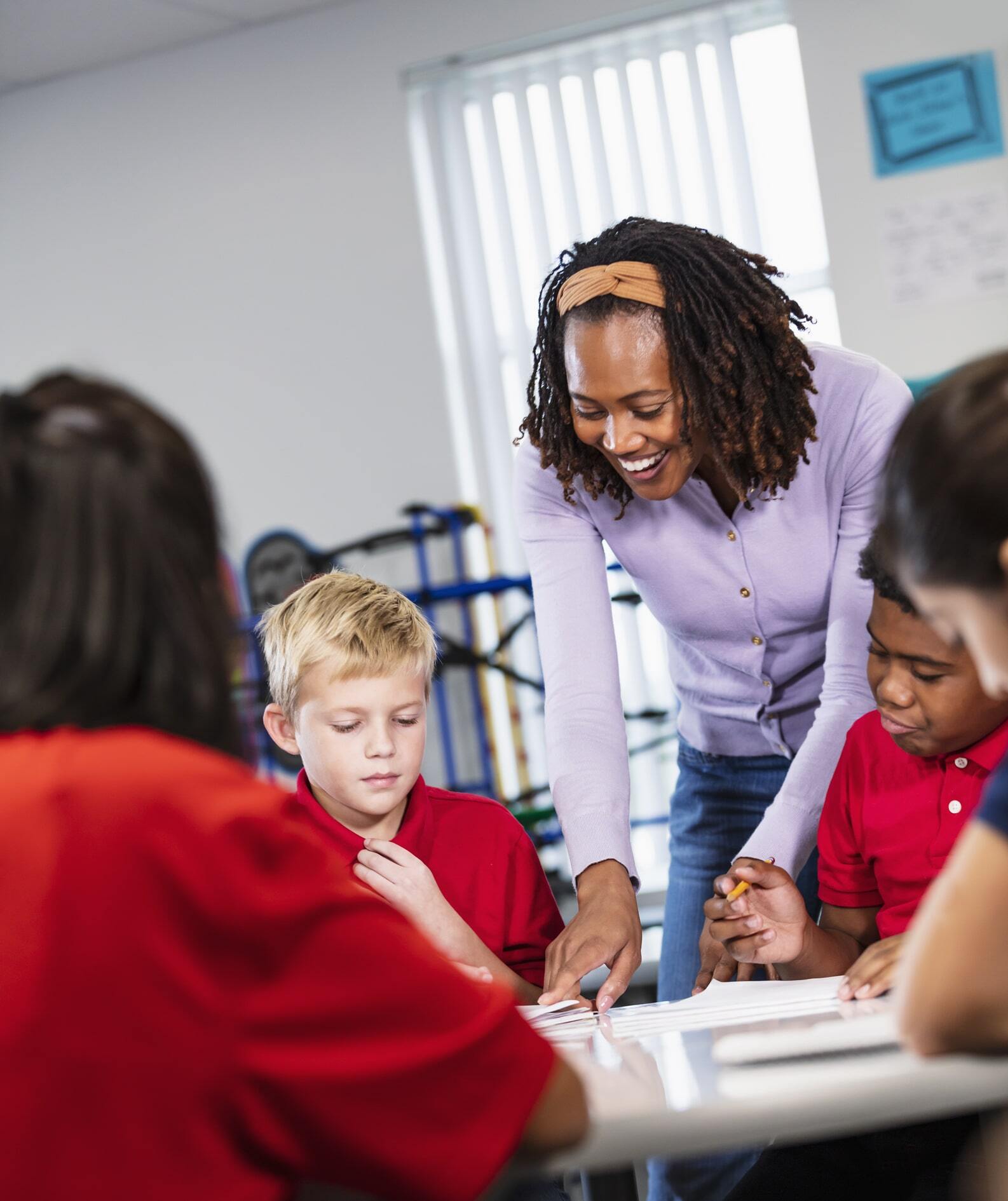 An African-American teacher helping a multiracial group of middle school students who are sitting at a table in a classroom writing in notebooks. The 11- and 12-year-old children are completing an assignment or taking a test
